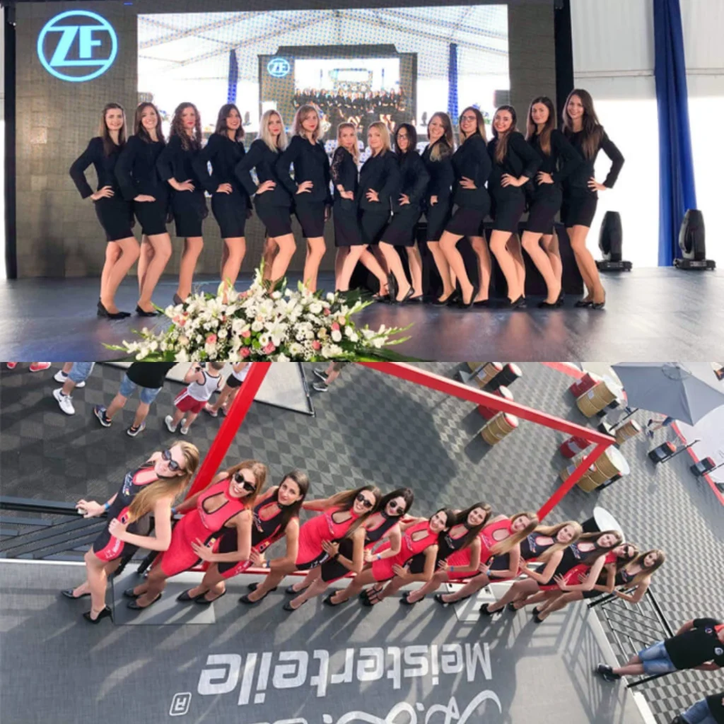 Fekete ruhás nők pózolnak a színpadon, a háttérben ZF logóval, virágkompozíció mellett. A közelben egy másik csoport vörös ruhás nő áll egy ferde emelvényen a Meisterfeier rendezvényen, ahol a fotósok megörökítik a pillanatot. Ideális rendezvényszervezés vagy hostess Budapest lehetőségek iránt érdeklődőknek. Mielőtt Elindulsz a Hotel Expo-ra – Mit Kell Tudnod az Eseményről?